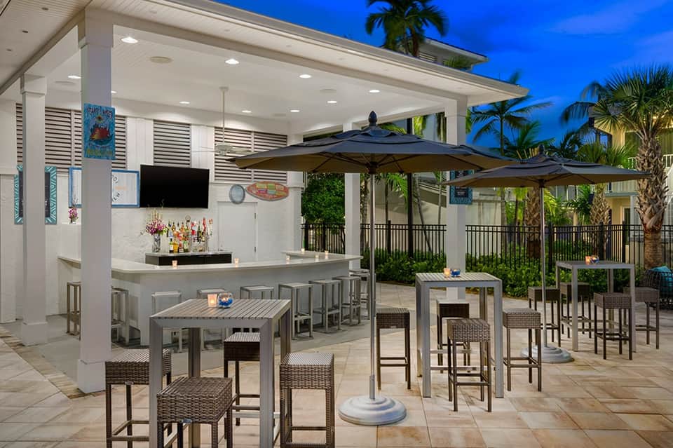 Beachfront tiki bar with white pergola, umbrellas, bar seating, and palm trees at dusk