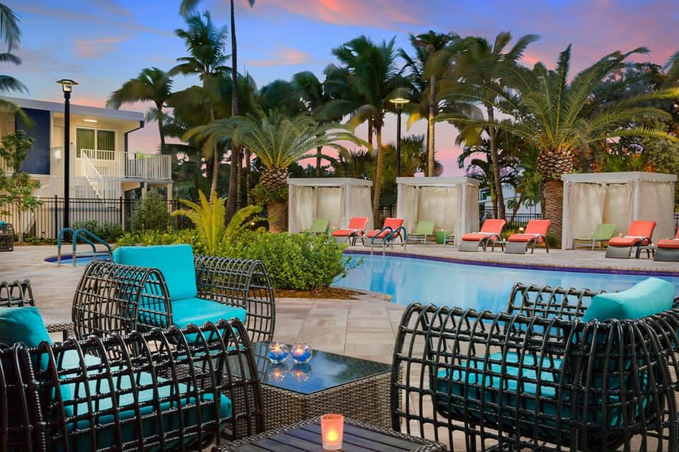 Resort pool deck at dusk with turquoise chairs, palm trees, and illuminated building in background