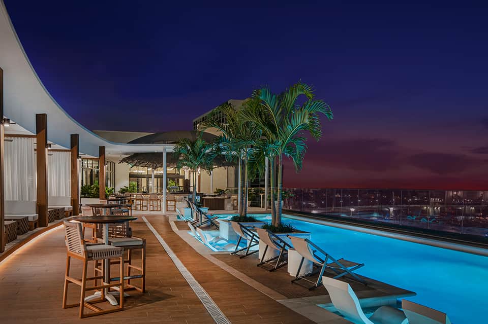 Elevated infinity pool overlooking cityscape at dusk with lounge chairs and palm trees