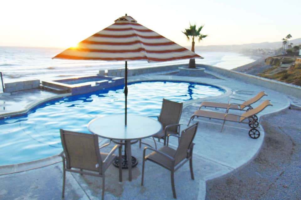 Oceanfront pool at sunset with striped umbrella, lounge chairs, and coastal cliffs