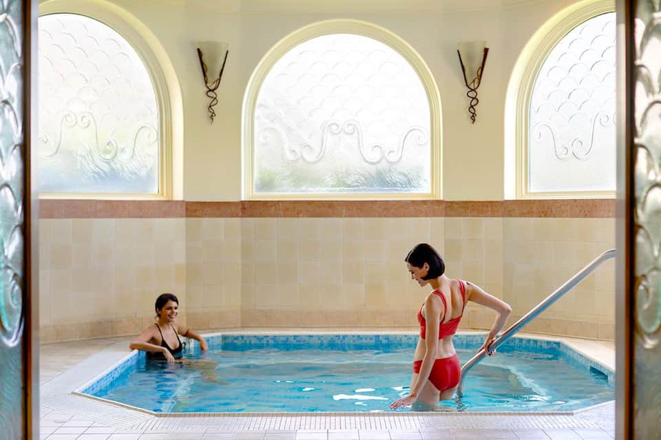 Indoor thermal spa pool with arched windows and two swimmers in a classical bathhouse setting
