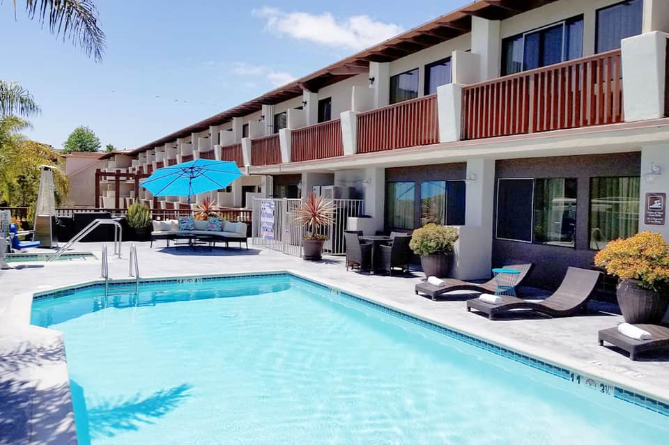 Modern hotel pool with lounge chairs, blue umbrella, mountain view, and multi-story residential building