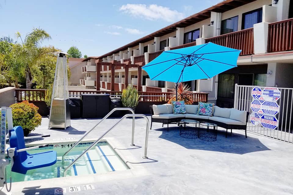 Resort pool deck with blue umbrella, seating area, and multi-story building in background