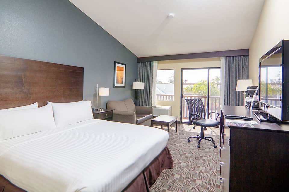 Hotel guest room with king bed, blue accent wall, desk, and balcony overlooking palm trees