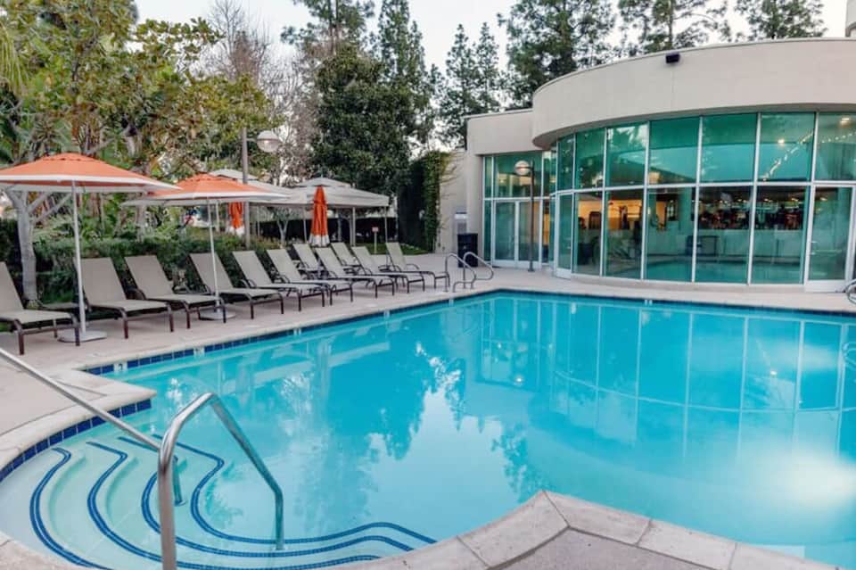 Resort pool with lounge chairs, umbrellas, and modern glass building with natural landscaping
