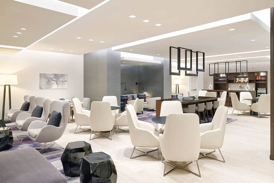 Modern hotel lounge with cream-colored curved chairs, bar area, and contemporary lighting fixtures.