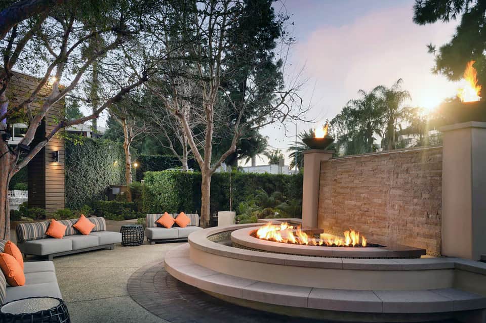 Evening patio with circular fire pit, lounge seating with orange pillows, illuminated torches, and mature trees