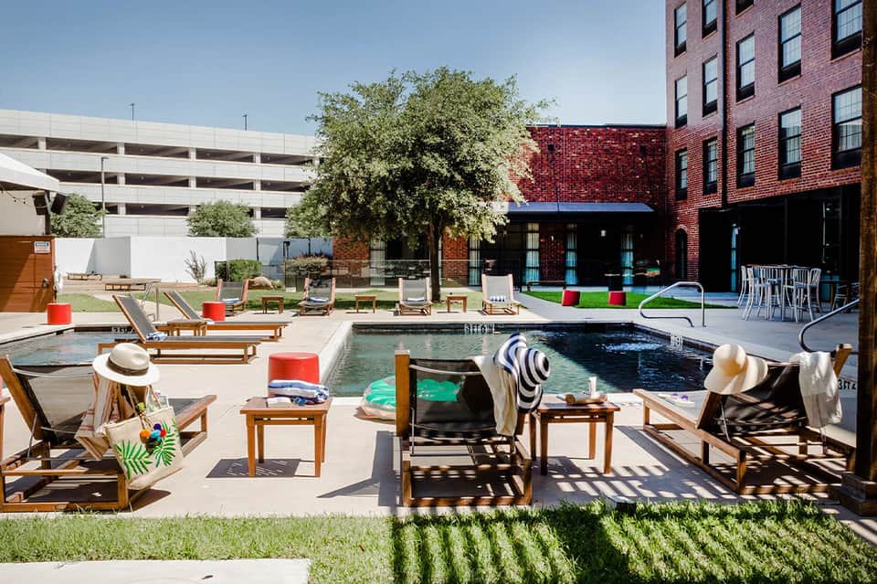 Urban resort courtyard with two pools, wooden lounge chairs, red accents, and brick building backdrop