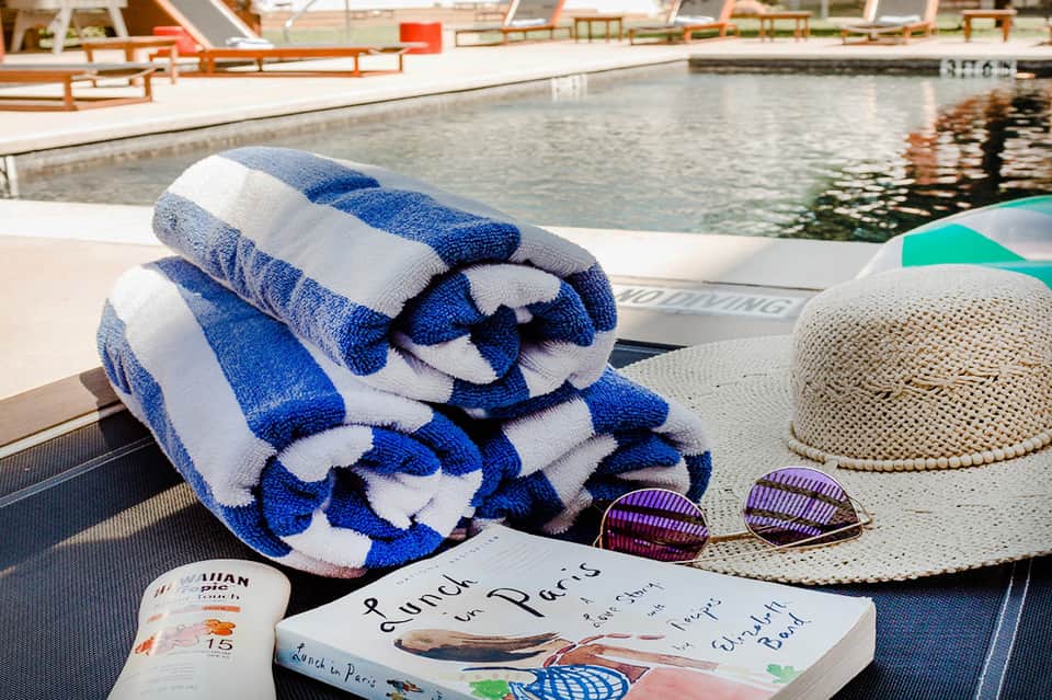 Poolside lounge setup with striped towels, sunscreen, hat, sunglasses, and book on chair.