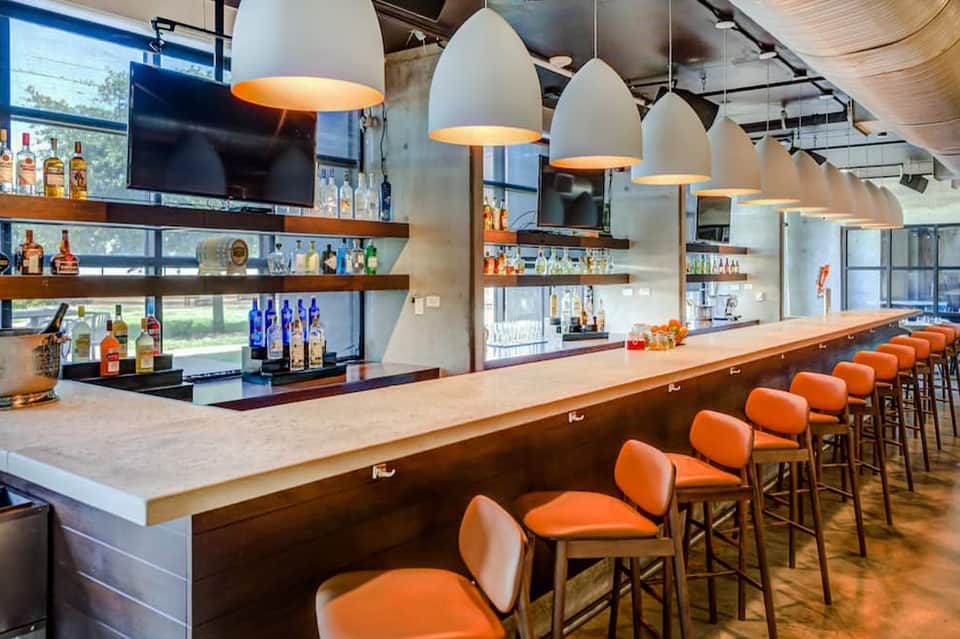 Modern bar interior with orange bar stools, pendant lights, and shelves of liquor bottles
