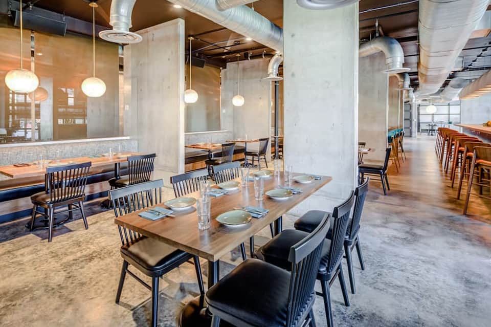 Modern industrial dining space with wooden tables, black chairs, concrete columns, and pendant lighting