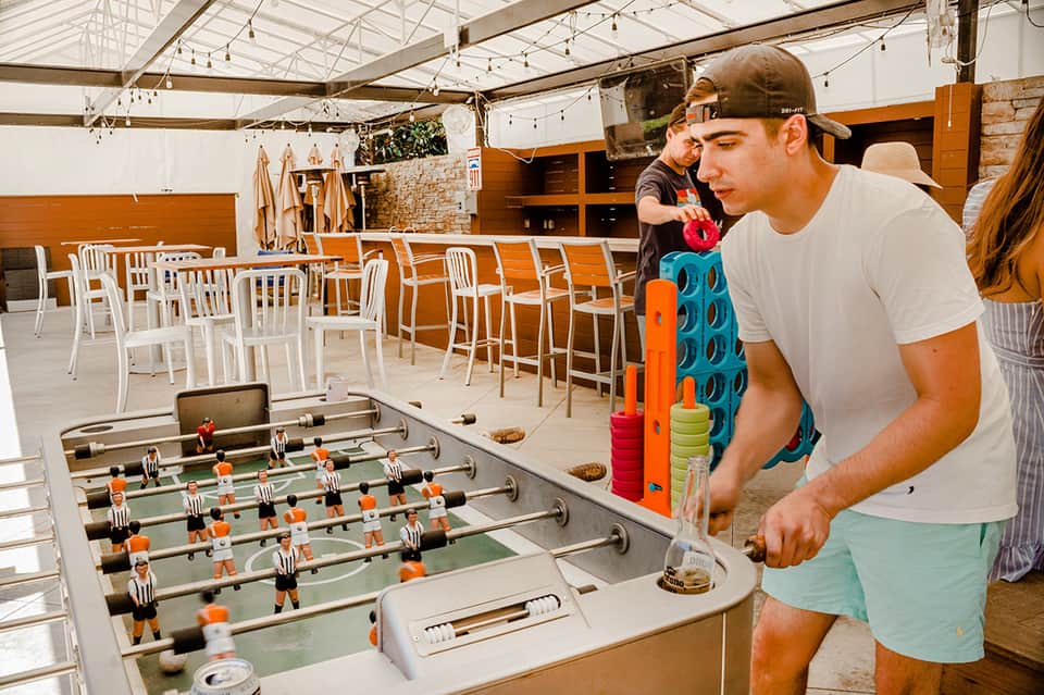 Man playing foosball in covered outdoor entertainment area with white chairs and wooden accents