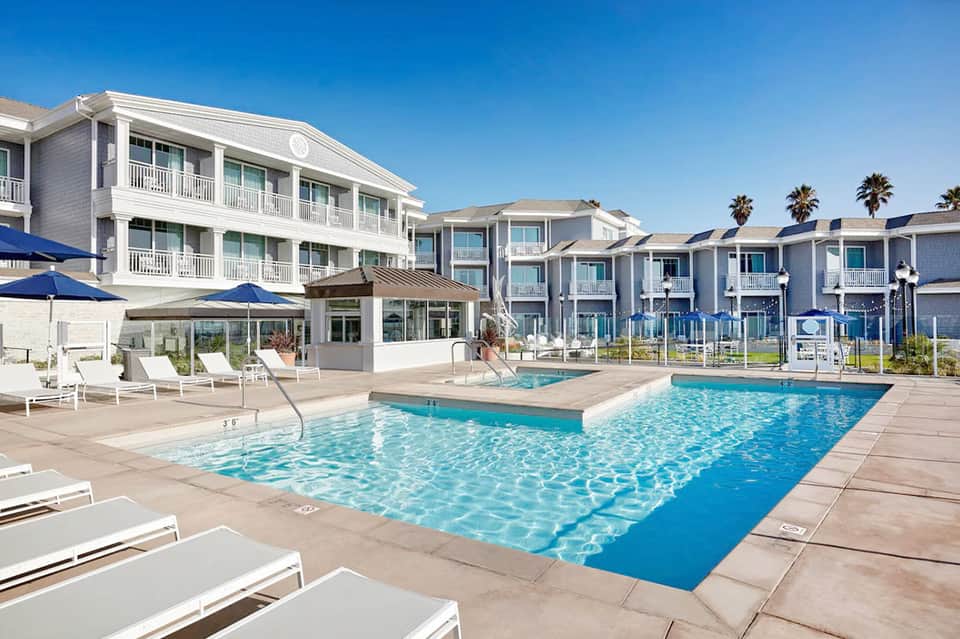 Large resort swimming pool with sun loungers, umbrellas, and beachfront hotel buildings under clear blue sky