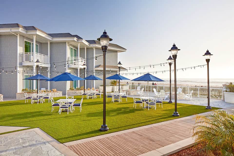 Seaside resort lawn with blue umbrellas, white furniture, and coastal mansion architecture with string lights.