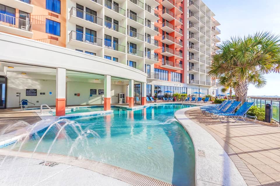 Resort pool with spa jets, colorful building facade, palm tree, lounge chairs, and covered patio area
