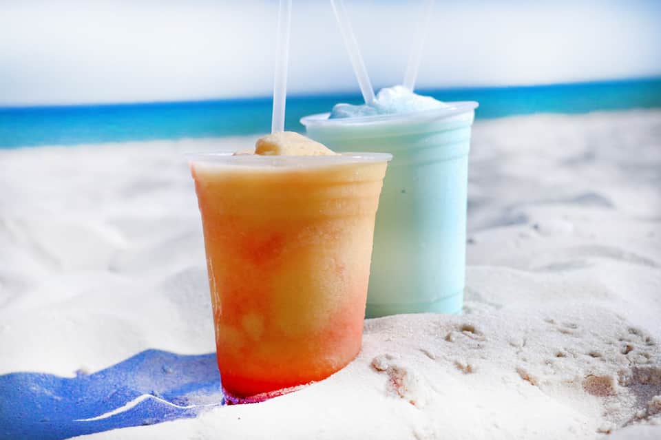 Colorful tropical cocktails in plastic cups on white sand beach with ocean backdrop