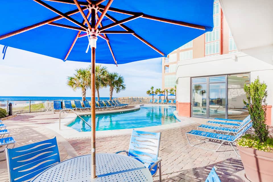 Beachfront pool with blue umbrellas, lounge chairs, and ocean view beside coral-colored building