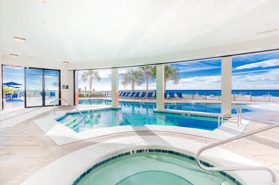 Indoor pool area with spa tub, overlooking ocean, palm trees and beach through large windows