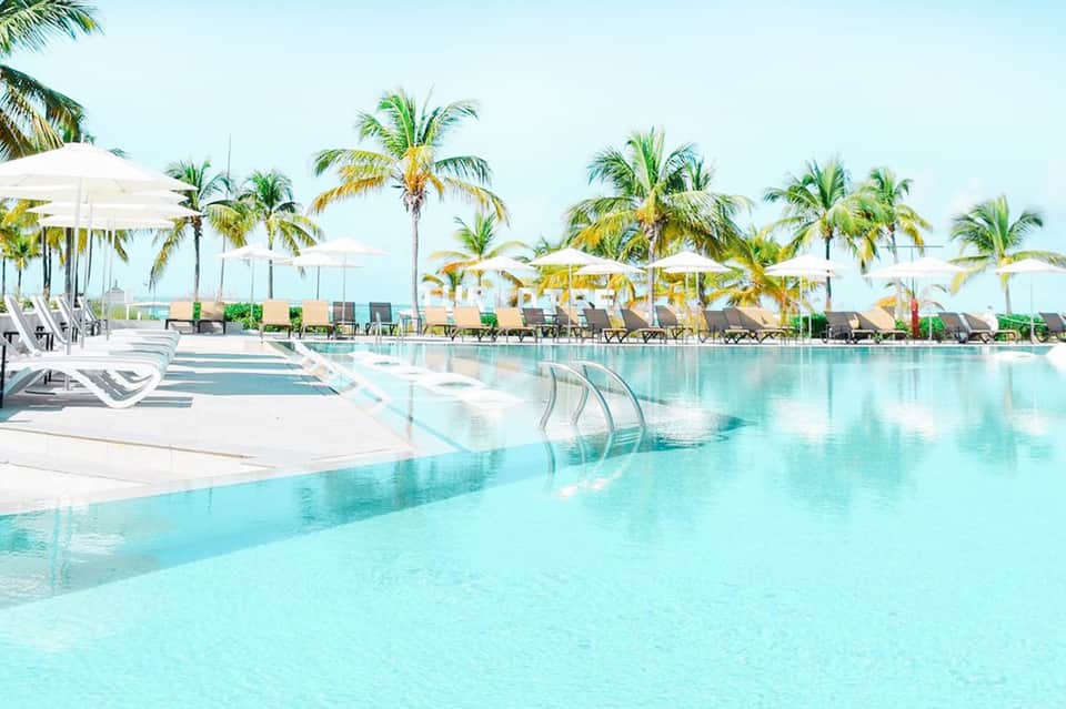 Turquoise resort pool with lounge chairs, white umbrellas, and palm trees in background