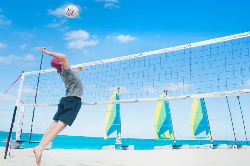 Beach volleyball player spiking ball with sailboats visible across turquoise water