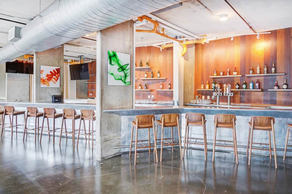 Contemporary bar interior with wooden seating, concrete walls, extensive bottle display, and industrial design elements