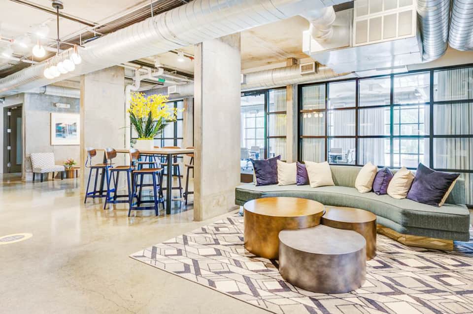 Modern hotel lobby with industrial ceiling, metallic round tables, green curved sofa, and bar seating area with natural light