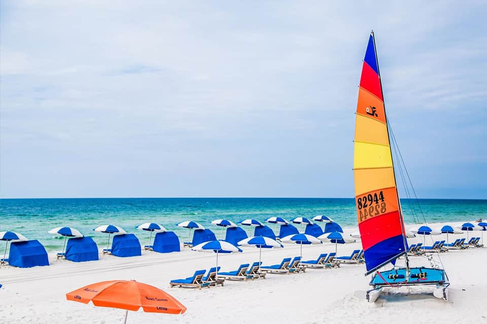 Colorful sailboat on white sand beach with blue umbrellas and loungers facing ocean