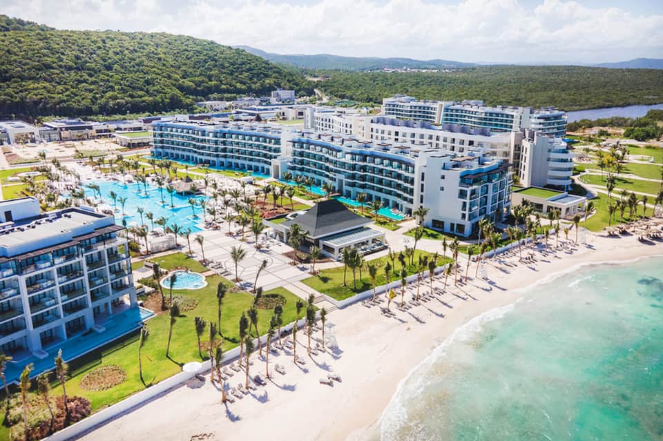 Aerial resort view with large pool complex, white buildings, beach loungers, and lush green surroundings