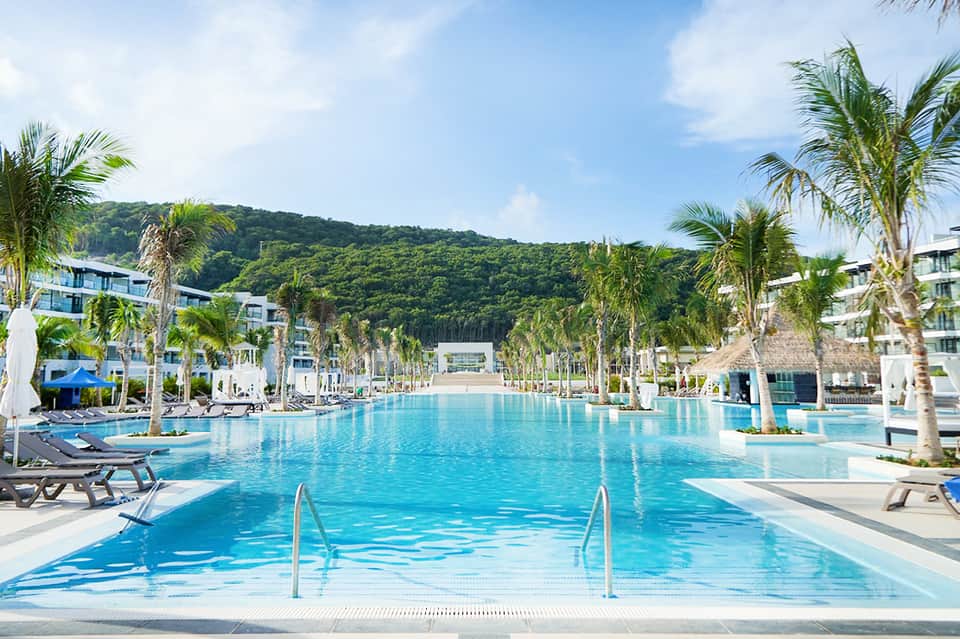 Resort infinity pool with palm trees, lounge chairs, and mountain views under clear blue sky
