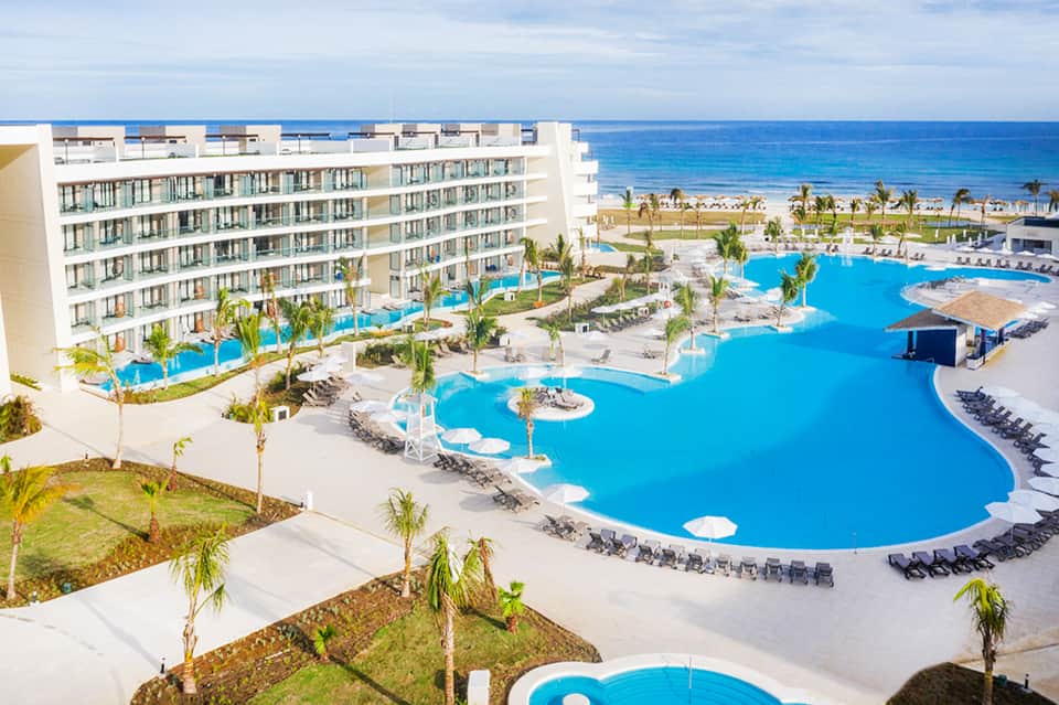 Beachfront resort with expansive curved pool, lounge chairs, white building, ocean and palm trees in background