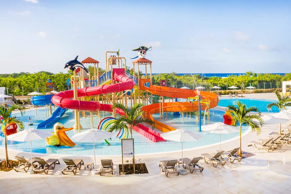 Colorful waterpark with red and blue water slides, splash pad, palm trees, and lounge chairs