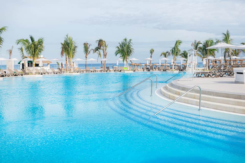 Resort-style pool with palm trees, lounge chairs, and beach in background