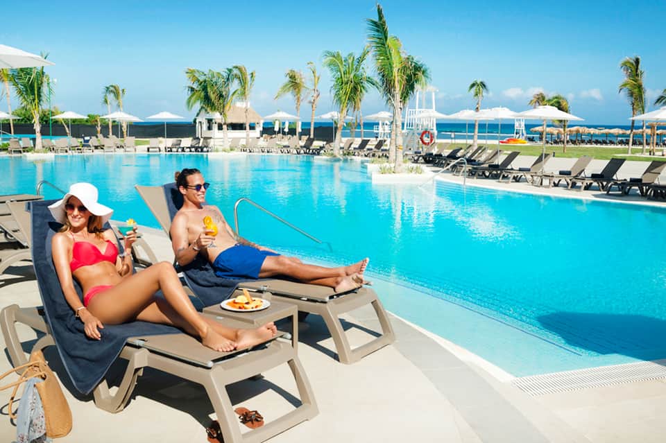 Two adults relaxing on lounge chairs by large resort pool with ocean and palm trees