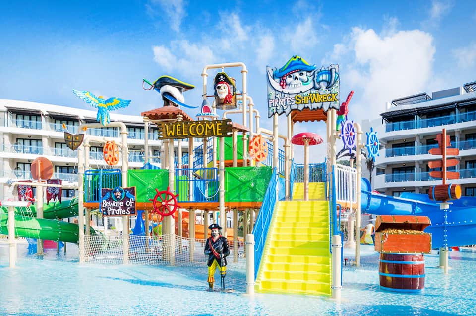 Pirate-themed water park playground with bright colors, slides, and shallow pool surrounded by resort buildings