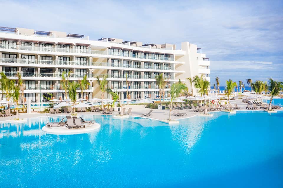 Large resort with white building and turquoise pool, palm trees, and ocean view backdrop