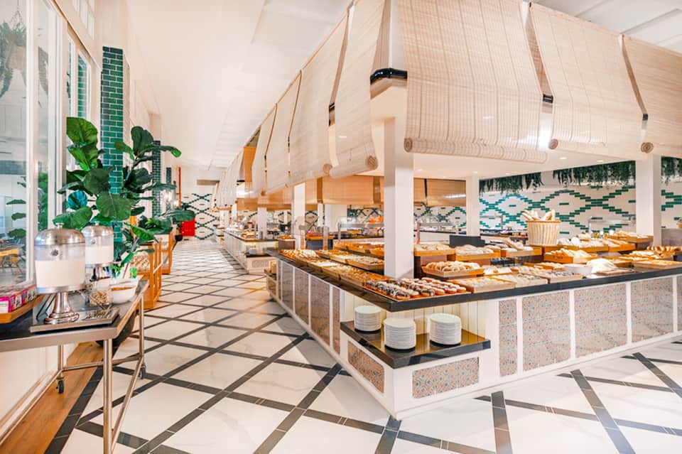 Modern resort dining hall with long counters, hanging mesh screens, tiled walls, and abundant natural light