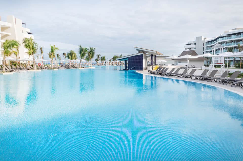 Large resort pool with crystal blue water, palm trees, and modern hotel buildings
