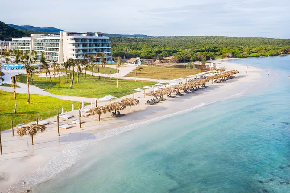 Beachfront resort with turquoise water, white sand, tiki umbrellas, and lush green grounds