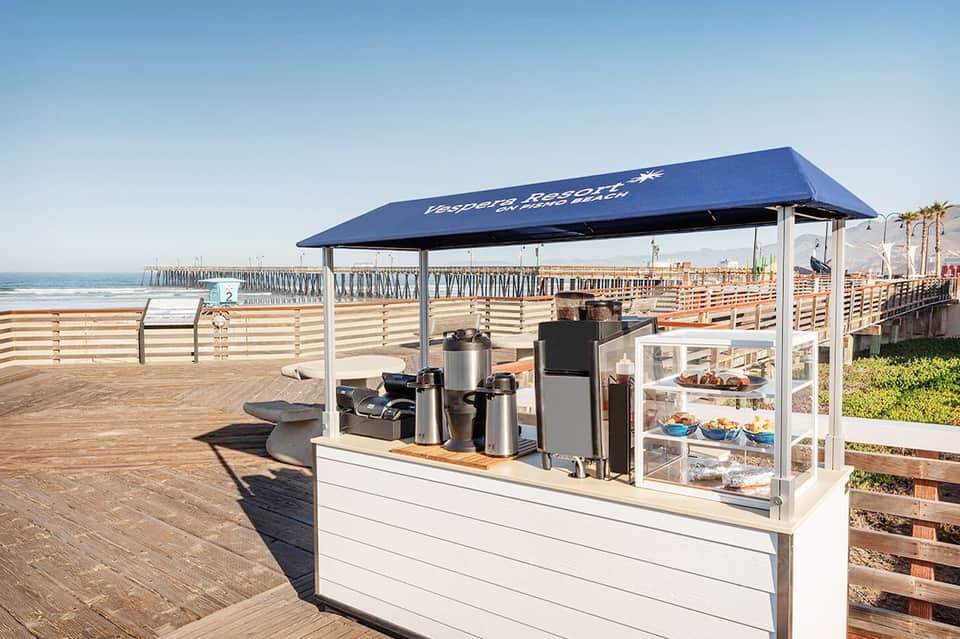 Beachside beverage stand with blue awning, pier and ocean view in background