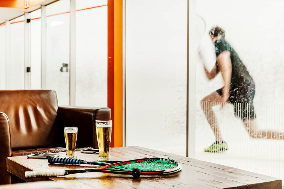 Indoor lounge with leather seating, tennis rackets, beers on wooden table, and squash player visible through glass