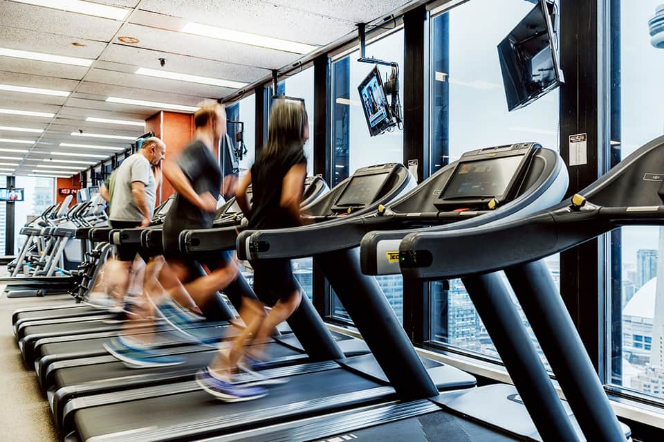 Multiple guests running on treadmills at modern gym with floor-to-ceiling windows and city views.