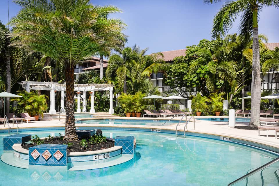 Resort pool with palm trees, white pergola structures, lounge chairs, and multi-level deck areas