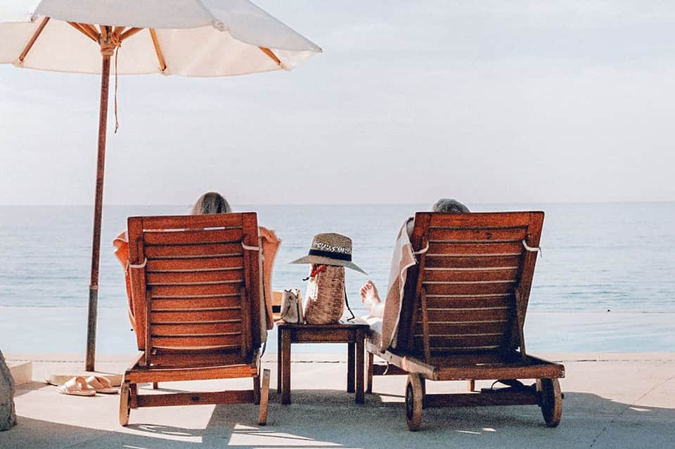 Couple in wooden lounge chairs under white umbrella relaxing on beach facing calm ocean