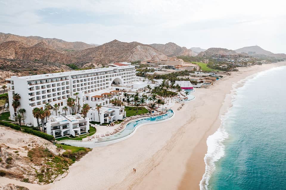 Modern white beachfront resort with curved pool, palm trees, mountains, and turquoise water in background