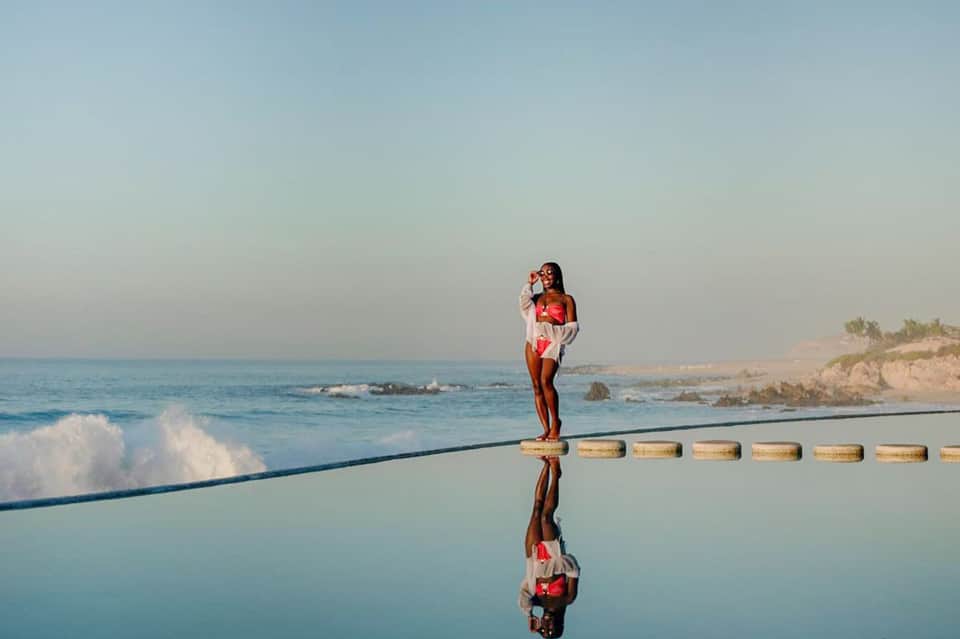 Woman in red outfit standing on wooden pier over infinity pool with ocean and beach horizon reflection