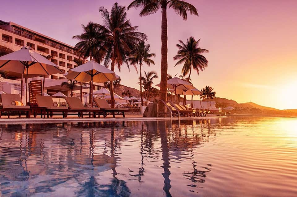Luxury beach resort at sunset with lounge chairs, umbrellas, palm trees and waterfront reflection