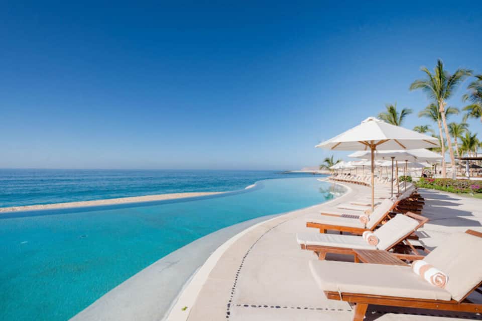 Luxury beachfront pool overlooking ocean with white lounge chairs, umbrellas, and palm trees