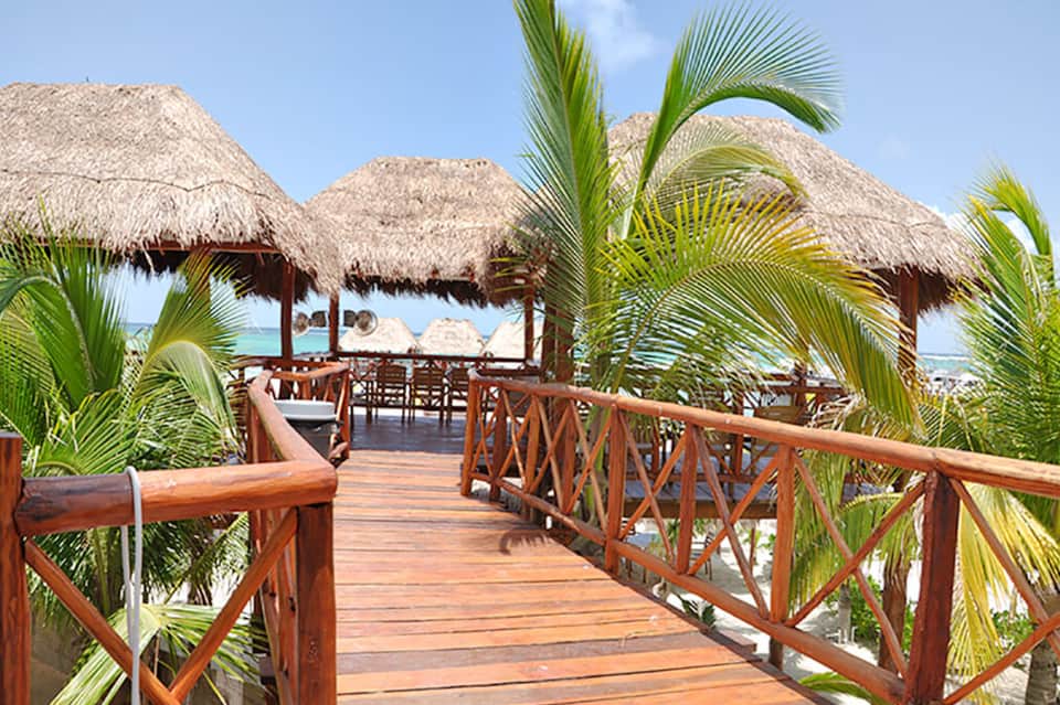 Wooden boardwalk with palm trees leading to thatched-roof beach cabanas overlooking turquoise ocean