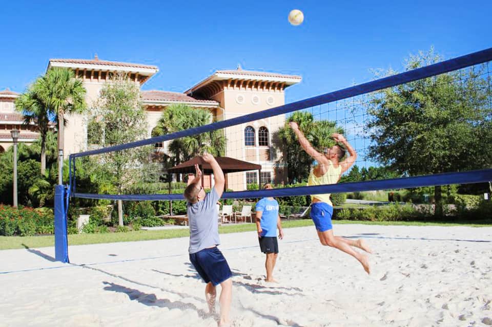 Three men playing beach volleyball with Spanish-style resort building in background under blue sky