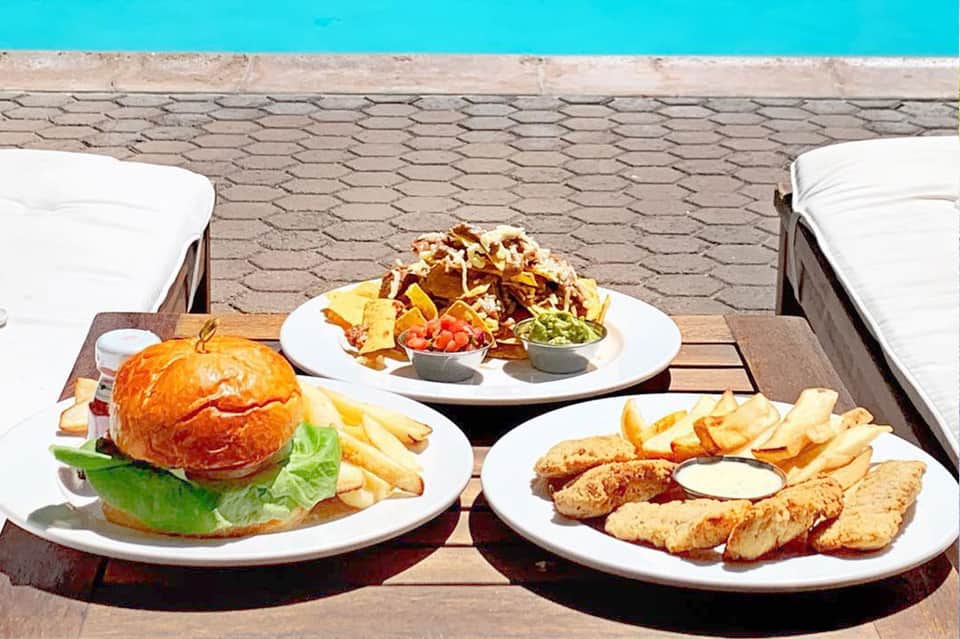 Poolside food service with burger, fried chicken strips, nachos, and condiments on table by turquoise pool